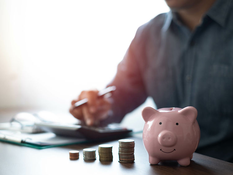 A piggy bank and stacks of coins in the foreground with a man on a laptop in the background