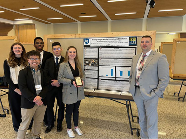 Penn State Altoona students Jazzmine McCauley, Ziwei (Will) Lin, Ray Ncube, Lam Vo, and Katelyn Kurtz standing next to their first-place poster, “Building Bridges with Pennsylvania State Police: An Assessment of Community Outreach,” and Trooper Joe Dunsmore, keynote speaker of the 2024 PACJE conference.  