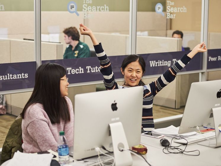 two students sitting together at computer stations, student at right smiles with arms raised joyfully