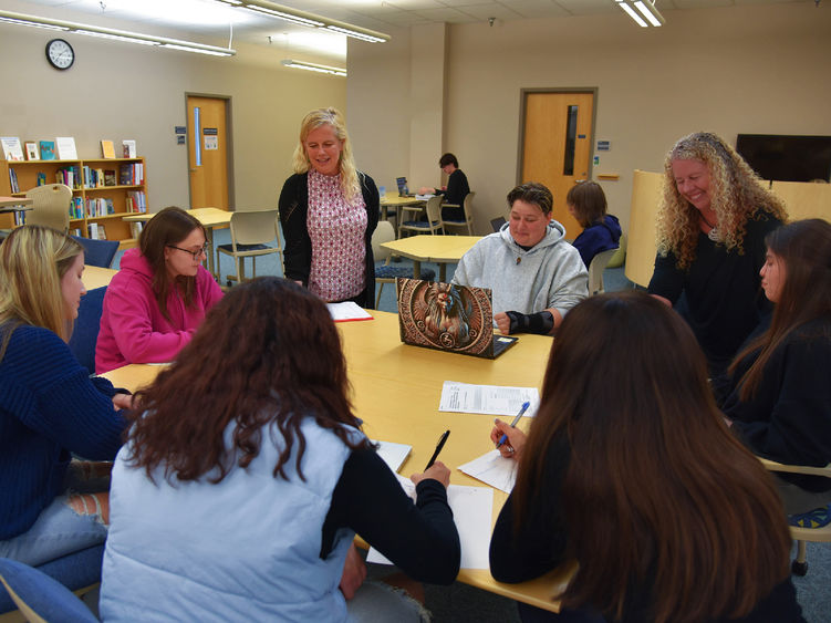 A group of students sits around a table in a library or classroom setting, engaged in discussion and writing. Two instructors stand with them, smiling and interacting. Bookshelves, chairs, and a clock are visible in the background, suggesting an academic or collaborative learning environment.