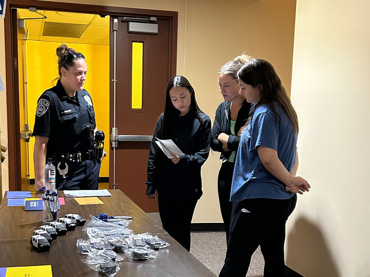 A police officer speaks with three women who are reading from a pamphlet at a table with promotional items in a hallway.