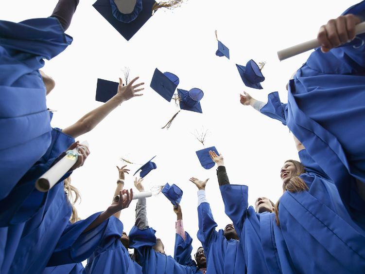 A group of students wearing blue graduation robes throw their caps in the air