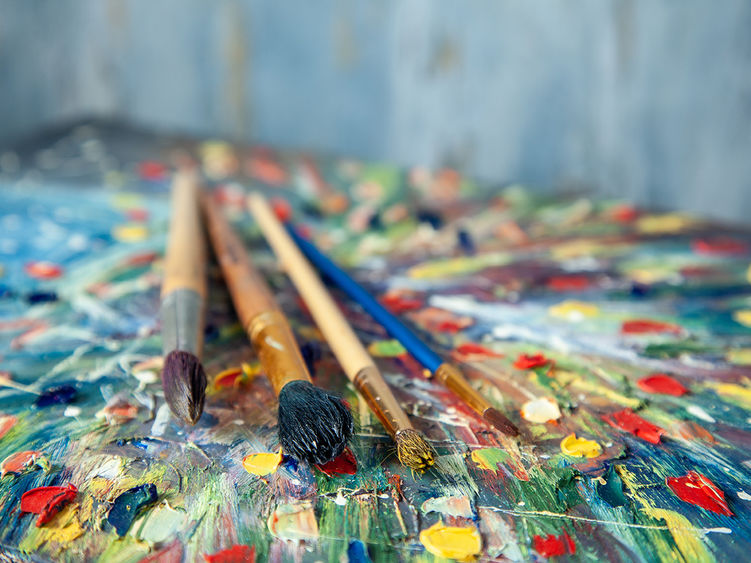 This close-up photo shows paintbrushes resting on a vibrant, abstract palette with thick dabs of colorful paint, evoking a lively artistic atmosphere.