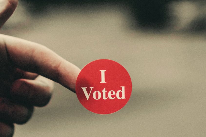closeup image of person's hand holding red "I Voted" sticker on fingertip