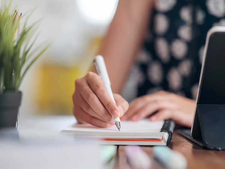 The hands of a woman writing on a notepad with a laptop computer sitting nearby