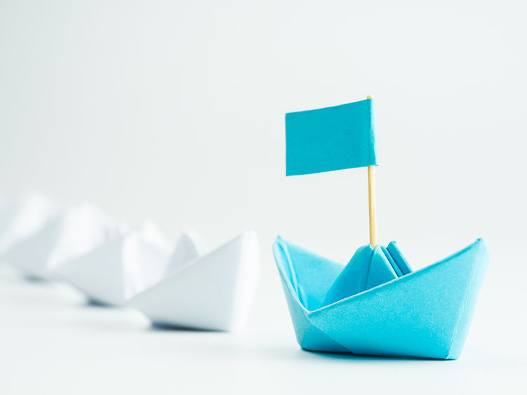 a blue paper sailboat leading other white paper sailboats